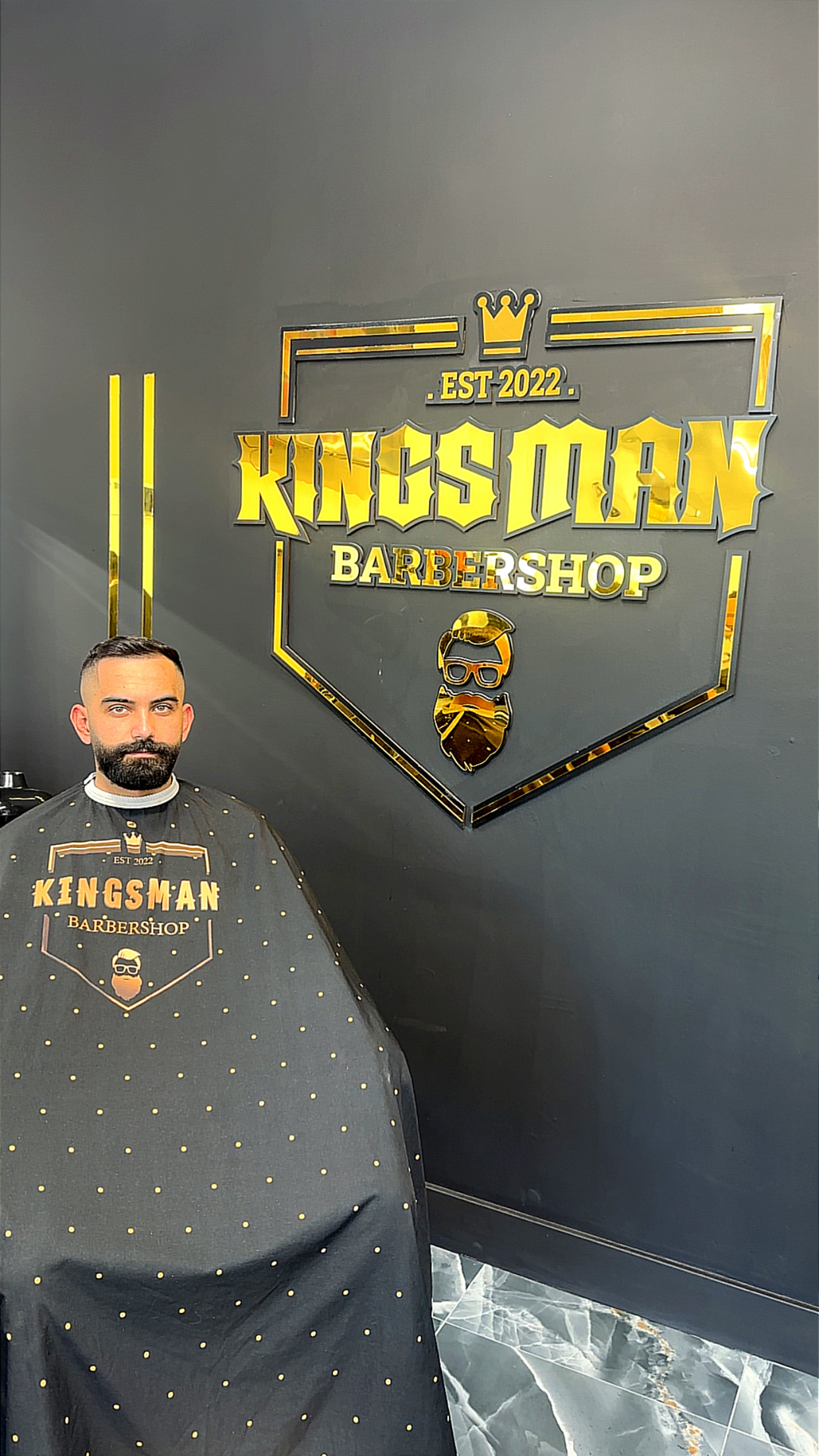 A client in the Kingsman cape in front of the shop's gold Est. 2022 logo wall