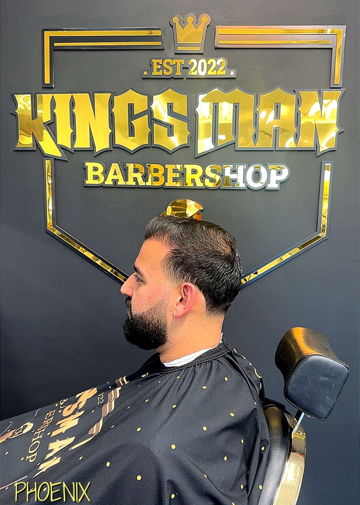 Low taper fade with beard, cut by Samer in front of the gold Kingsman logo wall